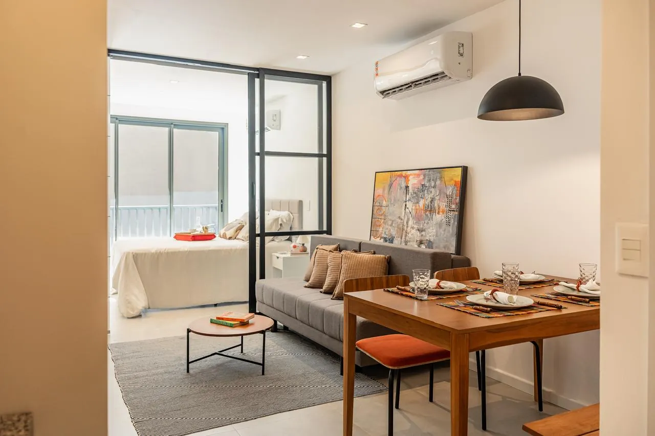 Foto real de um studio decorado do Glória Del'Art, com sala, mesa de jantar e quarto atrás da divisória de vidro