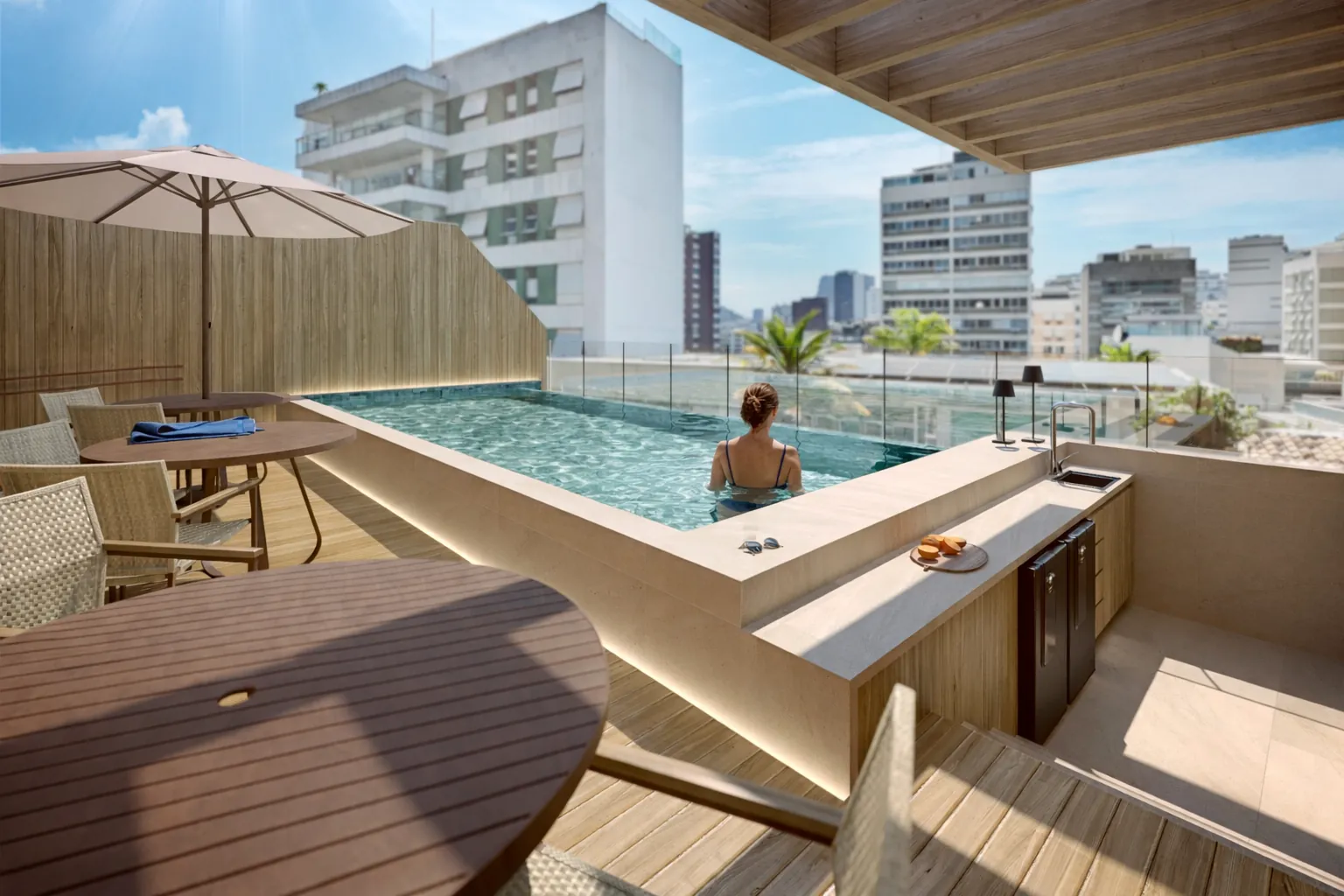 Piscina do rooftop com deck de madeira, mesa e os prédios do Leblon em volta
